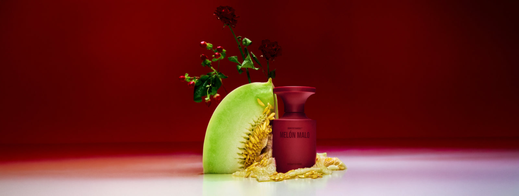 Melon Malo perfume bottle beside green melon and dark red flowers