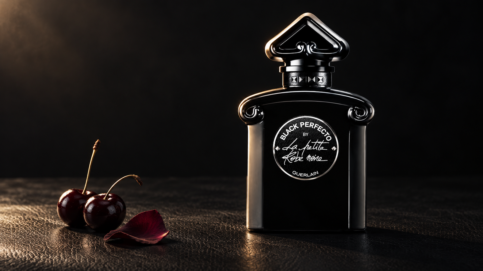 Guerlain Black Perfecto bottle beside two dark cherries and a rose petal on a black textured surface.