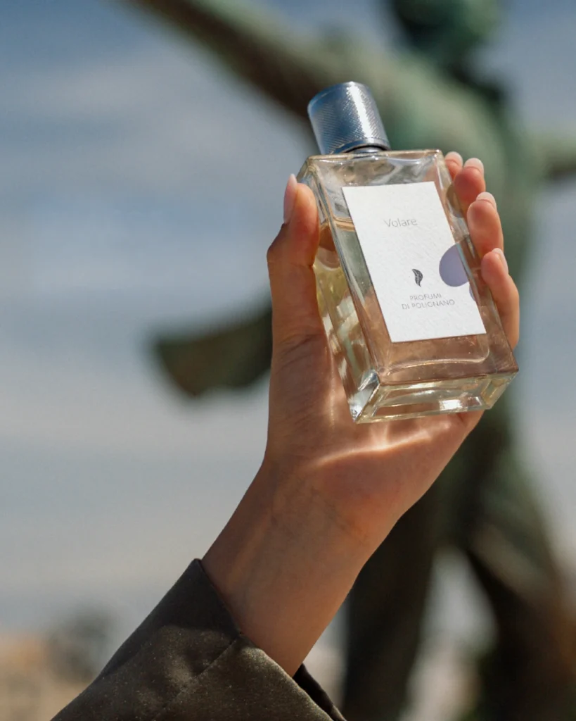 Hand holding a Volare perfume bottle outdoors against a softly blurred background.