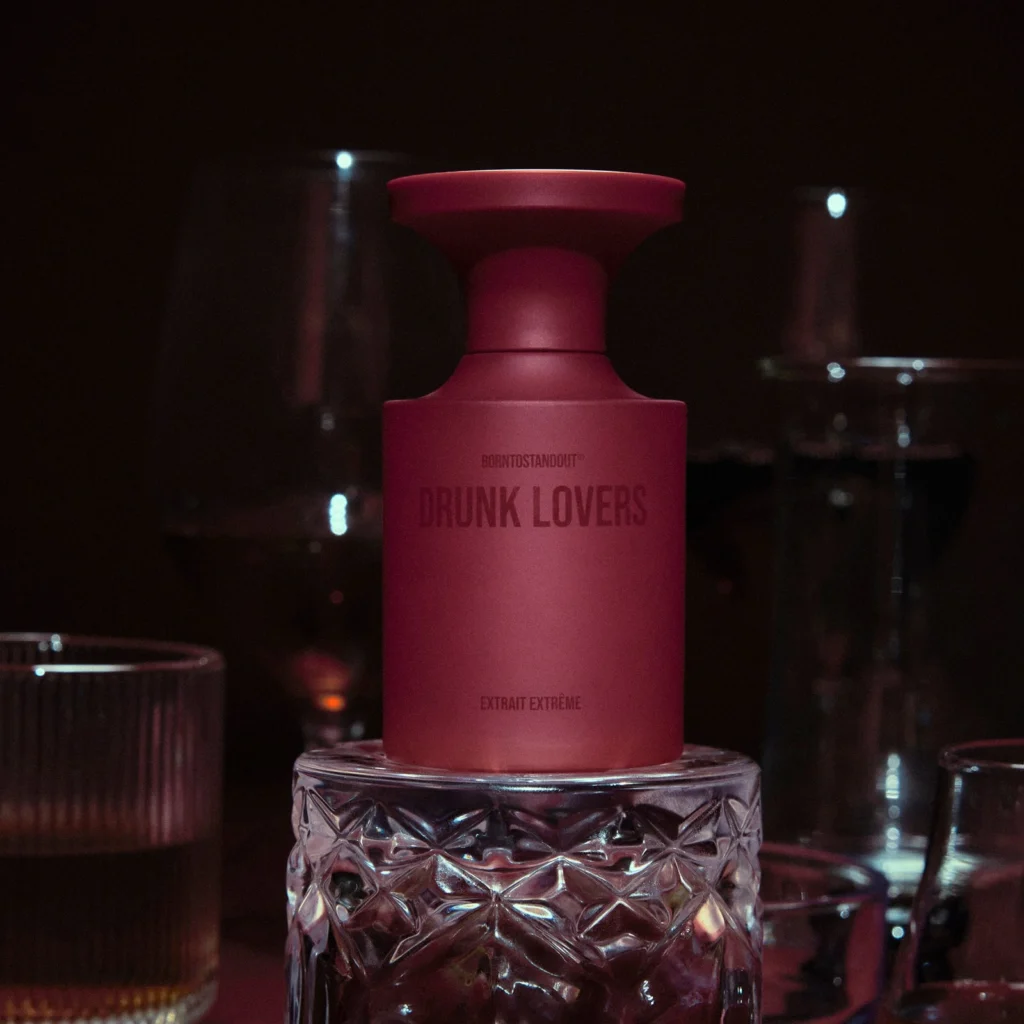 BORNTOSTANDOUT Drunk Lovers Extrait Extrême perfume bottle surrounded by drinking glasses in a dark setting