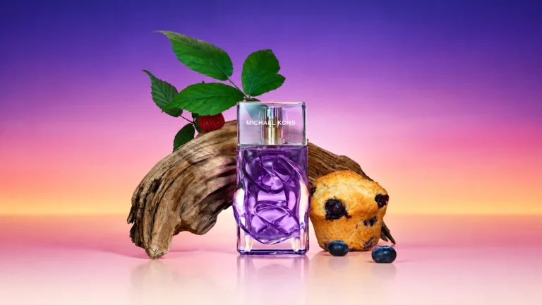 Michael Kors Violet Sunset Eau de Parfum surrounded by raspberries, blueberries, a muffin and driftwood against a purple sunset background.