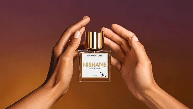 Nishane Hold Me Closer Extrait de Parfum framed by two hands against a warm burgundy and amber background.