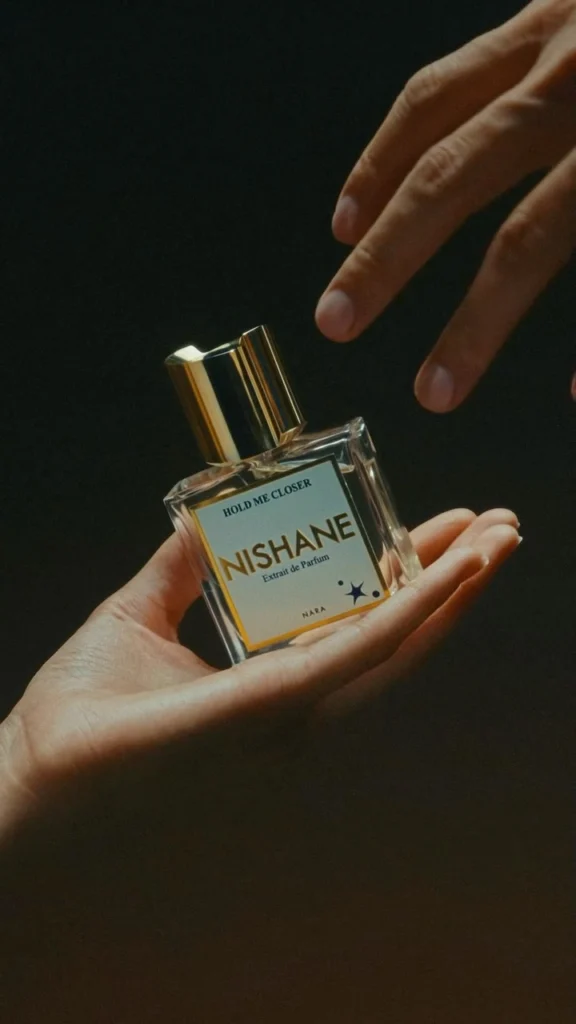 Nishane Hold Me Closer perfume bottle resting in an open hand as another hand reaches towards it against a dark background.