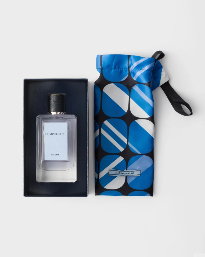 Prada Compulsion Eau de Parfum bottle displayed alongside its blue geometric-patterned satin pouch.